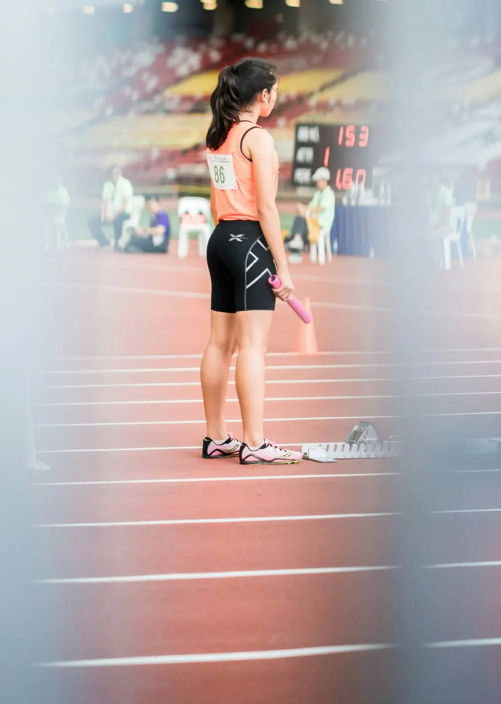 A relay runner stands alone on the track holding her baton, waiting for the race, seen past a blurred foreground rail.