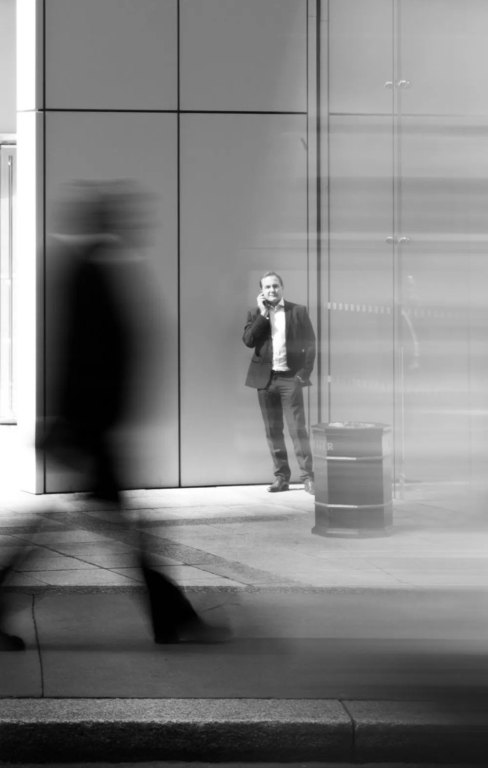 A man stands against a glass office wall, on the phone, while the people around him pass as silhouettes and motion blur.